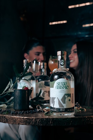 A wooden table displays a bottle labeled 'SEXO ABISMO', accompanied by a few symbolic objects like a mug with a raspberry and leaves, and a shiny ring. In the background, a blurred image of two people sits, suggesting a dimly lit, intimate setting with a convivial atmosphere.