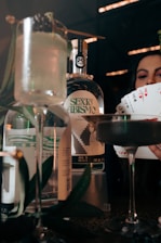 Bottles of gin with a label reading 'Sexto Abismo' are on display with a focused background, featuring a woman's hand holding a fan of playing cards, including aces and a joker. The scene is set with cocktail glasses, one containing a light-colored drink garnished with a twig or leaf.