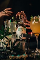 A collection of cocktail glasses and gin bottles are artfully arranged on a dark surface. Two hands with rings are gesturing near the bottles, which are labeled with a surrealist design. Garnishes like mint leaves and berries embellish the drinks, adding a touch of elegance.