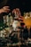 A collection of cocktail glasses and gin bottles are artfully arranged on a dark surface. Two hands with rings are gesturing near the bottles, which are labeled with a surrealist design. Garnishes like mint leaves and berries embellish the drinks, adding a touch of elegance.