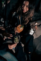 A woman with long hair and a bright smile is sitting in a dimly lit setting, holding a cocktail with garnishes. She is accompanied by another person holding a different drink. The scene conveys a social and relaxed atmosphere.