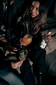 A woman with long hair and a bright smile is sitting in a dimly lit setting, holding a cocktail with garnishes. She is accompanied by another person holding a different drink. The scene conveys a social and relaxed atmosphere.