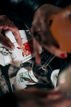 A hand holding playing cards featuring a Joker card, with an alcoholic beverage partially visible and a bottle labeled 'Sexto Abismo'. The scene suggests a casual gathering or game, with dim lighting adding a moody atmosphere.