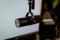 A professional black microphone is mounted on a stand against a blurry background, likely a part of a recording or broadcasting setup.