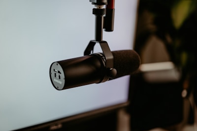 A professional black microphone is mounted on a stand against a blurry background, likely a part of a recording or broadcasting setup.
