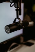 A black professional microphone mounted on a stand in a studio setting. The microphone is positioned in front of a blurred computer screen and keyboard, suggesting a recording or broadcasting environment.