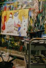 A vibrant art studio filled with colorful paintings and art supplies. In the foreground, a cart holds containers of paint and brushes. The background shows a large, abstract painting with vivid hues of red, yellow, and blue, along with other murals and paintings on the walls, creating a chaotic yet creative atmosphere.