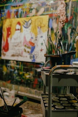 A vibrant art studio filled with colorful paintings and art supplies. In the foreground, a cart holds containers of paint and brushes. The background shows a large, abstract painting with vivid hues of red, yellow, and blue, along with other murals and paintings on the walls, creating a chaotic yet creative atmosphere.