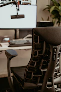 A modern office setup with a sleek ergonomic chair in the foreground. The chair features a complex support structure and black cushioning. Behind it, there is a computer desk with a large monitor displaying a bright screen and a microphone on an adjustable arm. A keyboard and phone can also be seen on the desk. Some greenery is present in the background, adding a natural element to the workspace.
