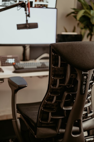 A modern office setup with a sleek ergonomic chair in the foreground. The chair features a complex support structure and black cushioning. Behind it, there is a computer desk with a large monitor displaying a bright screen and a microphone on an adjustable arm. A keyboard and phone can also be seen on the desk. Some greenery is present in the background, adding a natural element to the workspace.