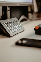 A desk setup featuring a monitor and a specialized keypad, perhaps for streaming or video editing. The scene also includes a keyboard and the edge of a smartphone, suggesting a tech-oriented workspace.