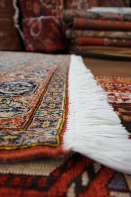 A close-up view of a richly patterned rug with intricate designs and a colorful border. The focus is on the edge with white fringes, and in the background, several folded rugs are visible.