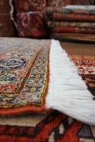 A close-up view of a richly patterned rug with intricate designs and a colorful border. The focus is on the edge with white fringes, and in the background, several folded rugs are visible.