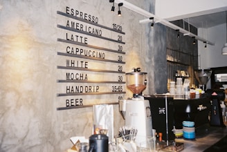 A modern cafe interior featuring a concrete wall with a menu listing various beverages including espresso, americano, latte, and beer, along with their prices. The counter displays coffee-making equipment such as a coffee grinder and espresso machine, surrounded by stacks of colorful cups and other accessories. Industrial-style pendant lighting hangs from the ceiling.