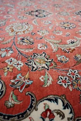 A detailed floral pattern with intricate designs featuring flowers and leaves in a variety of colors on a reddish-pink background. The artwork has a symmetrical and repeating motif typical of a classic Persian or oriental rug, showcasing fine craftsmanship and attention to detail.