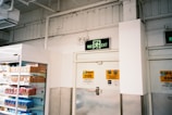 A supermarket aisle with a freezer containing packaged products to the left. Above is an exit sign with green emergency lighting. The walls are white with industrial piping, and there are safety notices near the door.