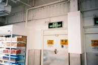 A supermarket aisle with a freezer containing packaged products to the left. Above is an exit sign with green emergency lighting. The walls are white with industrial piping, and there are safety notices near the door.