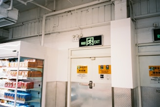 A supermarket aisle with a freezer containing packaged products to the left. Above is an exit sign with green emergency lighting. The walls are white with industrial piping, and there are safety notices near the door.