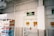 A supermarket aisle with a freezer containing packaged products to the left. Above is an exit sign with green emergency lighting. The walls are white with industrial piping, and there are safety notices near the door.