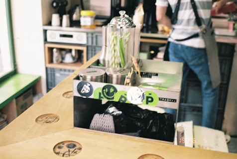 A casual indoor scene featuring a countertop with a box labeled for trash disposal. On top of the box are metal canisters and a glass container with straws. The background includes shelves with various items like jars and kitchen equipment. A person wearing a striped shirt and satchel is partially visible.