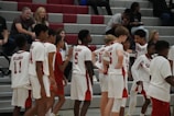 A group of young basketball players in uniforms is gathered on a court. They are dressed in white jerseys with red details. There are spectators seated in the background on bleachers, casually watching the scene.