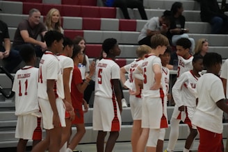 A group of young basketball players in uniforms is gathered on a court. They are dressed in white jerseys with red details. There are spectators seated in the background on bleachers, casually watching the scene.