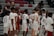 A group of young basketball players in uniforms is gathered on a court. They are dressed in white jerseys with red details. There are spectators seated in the background on bleachers, casually watching the scene.