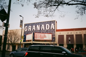 A theater marquee displays upcoming performances, with trees and a street in the foreground. The marquee reads 'GRANADA' with a list of artists and dates. Cars and people are visible near the theater. The sky is clear, and there are some bare tree branches overhead.