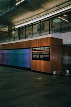A modern interior space with a glass-paneled upper level, featuring a large wall-mounted digital display and a wooden-paneled wall with signage for the Bill & Melinda Gates Center. The floor is made of dark stone tiles, and the environment has a clean, professional atmosphere.