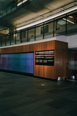 A modern interior space with a glass-paneled upper level, featuring a large wall-mounted digital display and a wooden-paneled wall with signage for the Bill & Melinda Gates Center. The floor is made of dark stone tiles, and the environment has a clean, professional atmosphere.