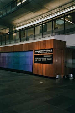 A modern interior space with a glass-paneled upper level, featuring a large wall-mounted digital display and a wooden-paneled wall with signage for the Bill & Melinda Gates Center. The floor is made of dark stone tiles, and the environment has a clean, professional atmosphere.