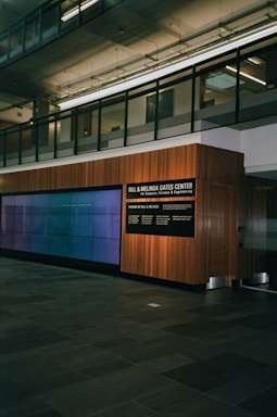 A modern interior space with a glass-paneled upper level, featuring a large wall-mounted digital display and a wooden-paneled wall with signage for the Bill & Melinda Gates Center. The floor is made of dark stone tiles, and the environment has a clean, professional atmosphere.
