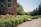 A campus-like environment with a large brick building featuring tall windows. A path leads through well-maintained gardens filled with colorful roses and green hedges. Trees line the area, providing a serene and natural backdrop.