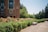 A campus-like environment with a large brick building featuring tall windows. A path leads through well-maintained gardens filled with colorful roses and green hedges. Trees line the area, providing a serene and natural backdrop.