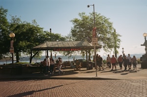 People are walking and gathering near an outdoor market area with a metal-roofed pavilion. The signage reads 'Pike Place Market' in red letters. Trees provide shade and a vibrant green backdrop. Other elements include street lamps, a one-way sign, and cobblestoned pavement. The sky is clear and blue.