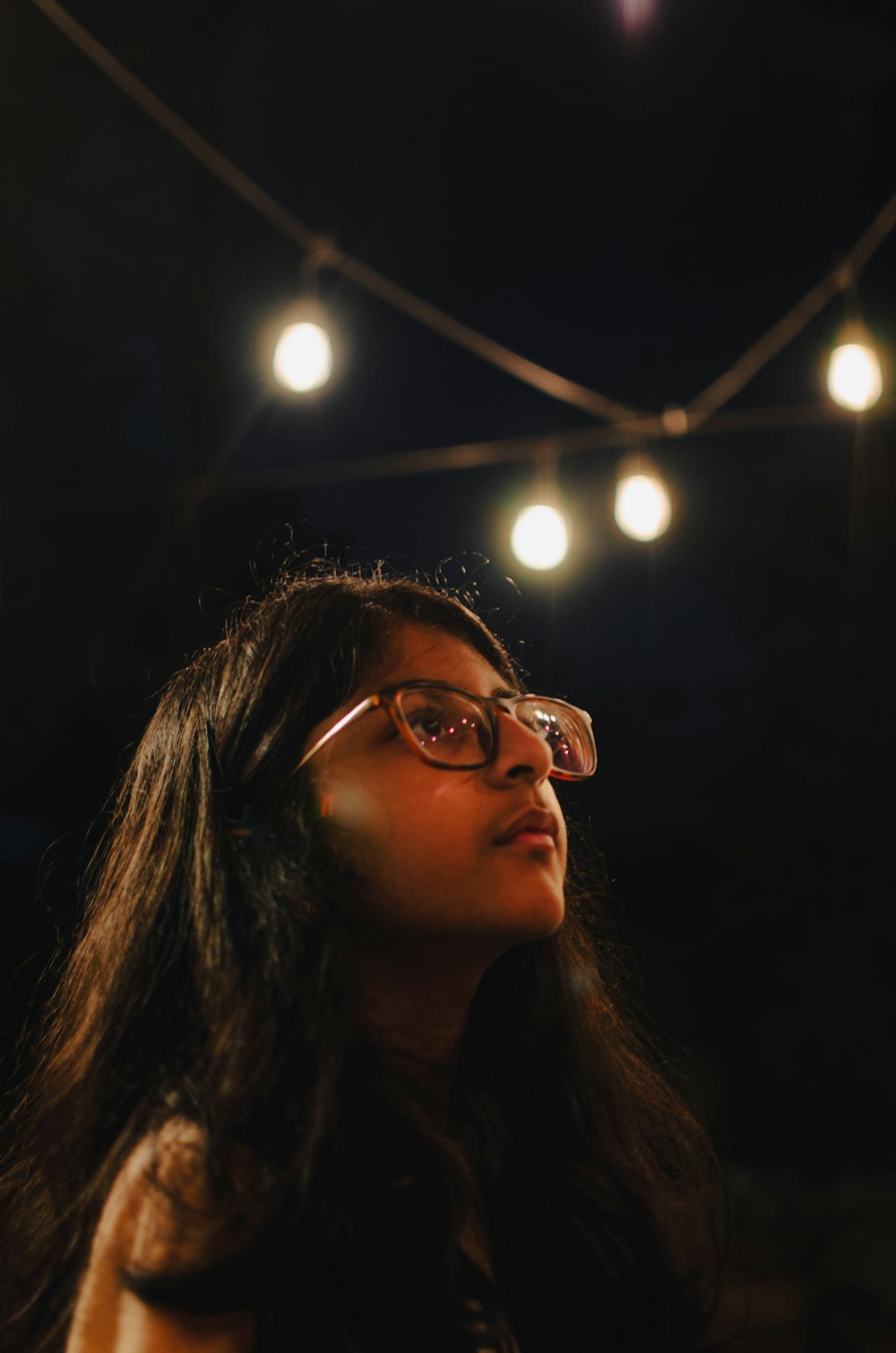 a woman wearing glasses looking up at a string of lights