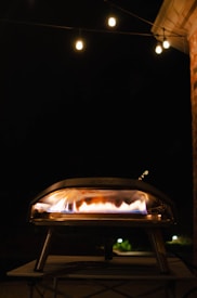 A pizza oven with visible flames is set against a dark night sky. Warm string lights hang above, providing a cozy outdoor ambiance.
