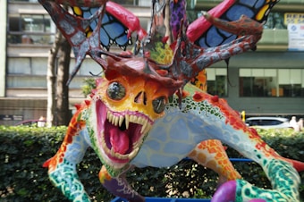 A colorful, fantastical creature with vibrant patterns and textures stands prominently. The creature has a wide-open mouth displaying large, sharp teeth, and its skin features a mix of bright colors including red, orange, blue, green, and yellow. The body is covered in scales and spots with wings that resemble butterfly wings. It seems to be situated outdoors, with foliage and urban structures in the background.