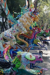 A vibrant collection of colorful, fantastical creatures with intricate patterns and designs, prominently featuring a large, multicolored lion-like figure with antlers. The setting appears to be an outdoor space with trees and paved paths, where these sculptures are displayed on platforms with wheels.