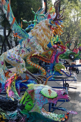 A vibrant collection of colorful, fantastical creatures with intricate patterns and designs, prominently featuring a large, multicolored lion-like figure with antlers. The setting appears to be an outdoor space with trees and paved paths, where these sculptures are displayed on platforms with wheels.