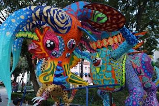 A vibrant, multicolored sculpture depicting fantastical creatures, each adorned with intricate patterns and designs. The creatures have exaggerated features like large eyes and swirling horns, combining elements of various animals and imaginary beings. The structure is set outdoors against a backdrop of trees and a building facade.
