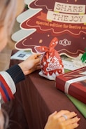 A person holds a red gift bag with 'Merry Christmas' written on it, adorned with white holiday-themed designs. In the background, there is a cardboard cutout in the shape of a Christmas tree with the message 'Share the Joy: special edition for festive moments.' The table is covered with a brown cloth, and nearby is a gift box wrapped with a ribbon.