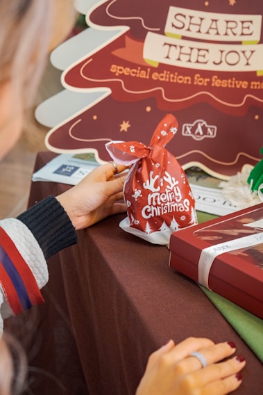 A person holds a red gift bag with 'Merry Christmas' written on it, adorned with white holiday-themed designs. In the background, there is a cardboard cutout in the shape of a Christmas tree with the message 'Share the Joy: special edition for festive moments.' The table is covered with a brown cloth, and nearby is a gift box wrapped with a ribbon.