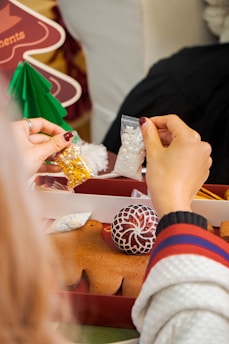 Hands holding small packets of decorative candies, possibly for decorating cookies or gingerbread. Visible gingerbread shaped as a person and other holiday-themed decorations are present.
