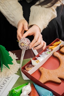 Hands are delicately handling a decorative ball ornament, surrounded by festive materials including gingerbread cookies, colorful icing bags, and crafting supplies. A small tree, various decorative elements like ribbons and shimmering powders are also present.