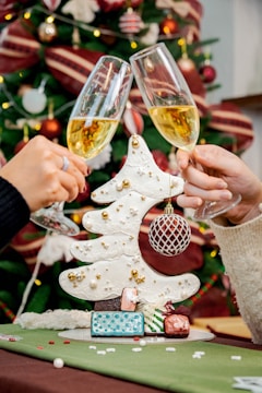 Two hands raise champagne glasses in a toast in front of a decorated Christmas tree. A white, stylized Christmas tree centerpiece adorned with gold accents and a hanging ornament is surrounded by small, colorful gifts.