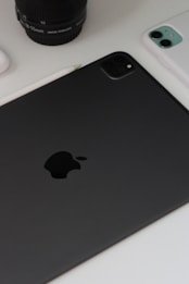 A close-up view of technology devices including a black tablet with a prominent logo, a white smartphone, a camera lens, and a stylus on a white surface.