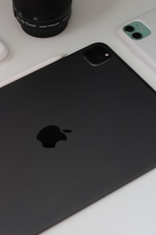 A close-up view of technology devices including a black tablet with a prominent logo, a white smartphone, a camera lens, and a stylus on a white surface.