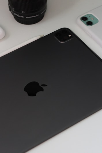 A close-up view of technology devices including a black tablet with a prominent logo, a white smartphone, a camera lens, and a stylus on a white surface.