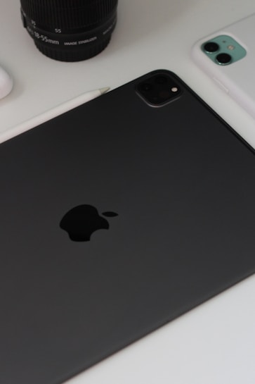 A close-up view of technology devices including a black tablet with a prominent logo, a white smartphone, a camera lens, and a stylus on a white surface.